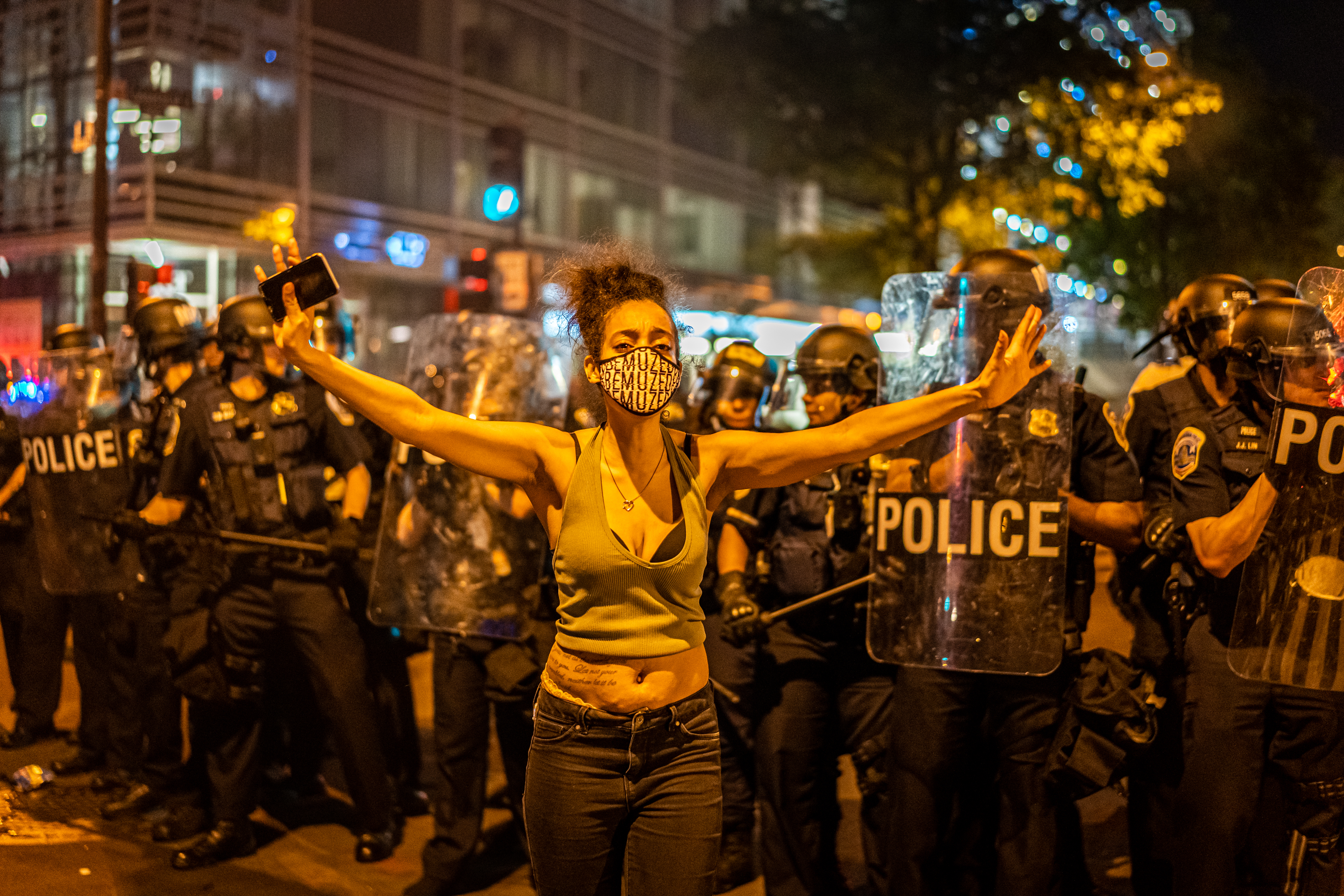 A woman holds her arms open, facing away from police holding shields.