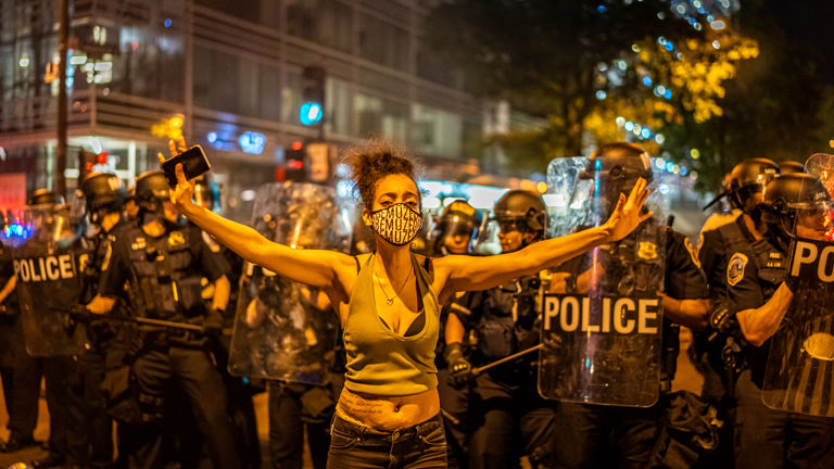 A woman holds her arms open, facing away from police holding shields.