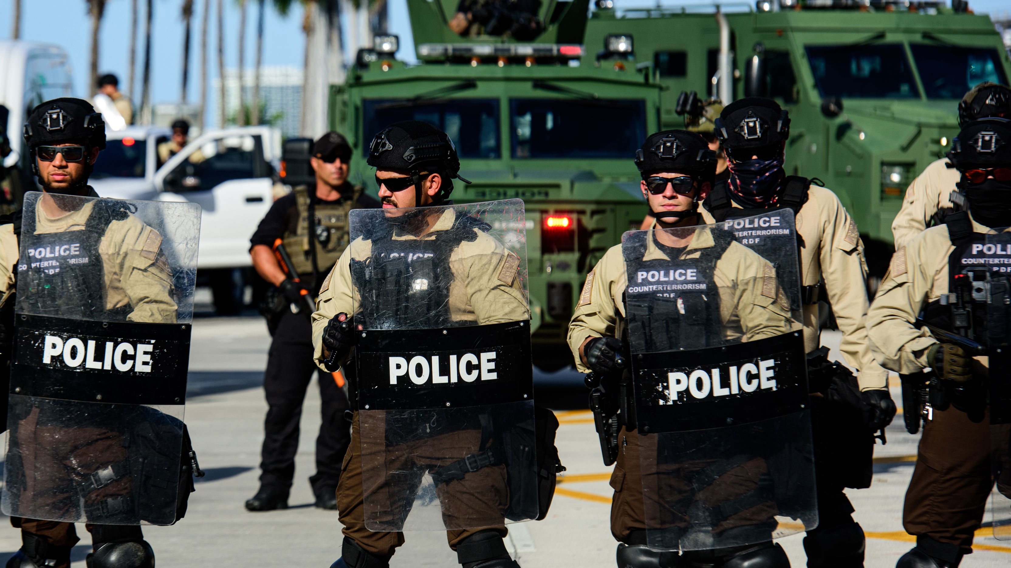 Several police officers in riot gear stand in the foreground. In the background, more officers drive green militarized trucks..