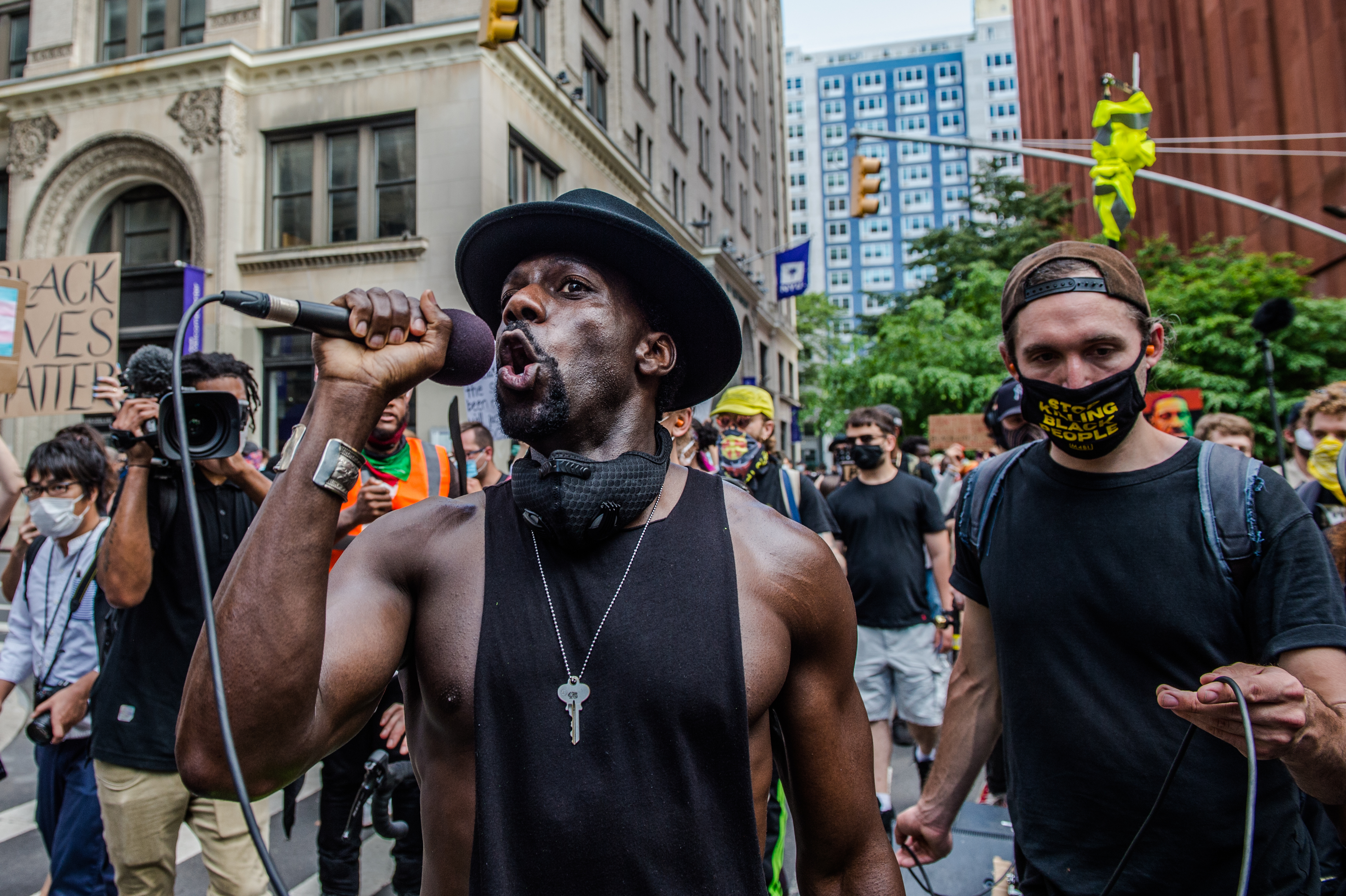 A man speaks into a microphone; a protestor in the background holds a "Black Lives Matter" sign
