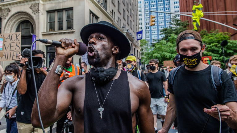 A man speaks into a microphone; a protestor in the background holds a "Black Lives Matter" sign